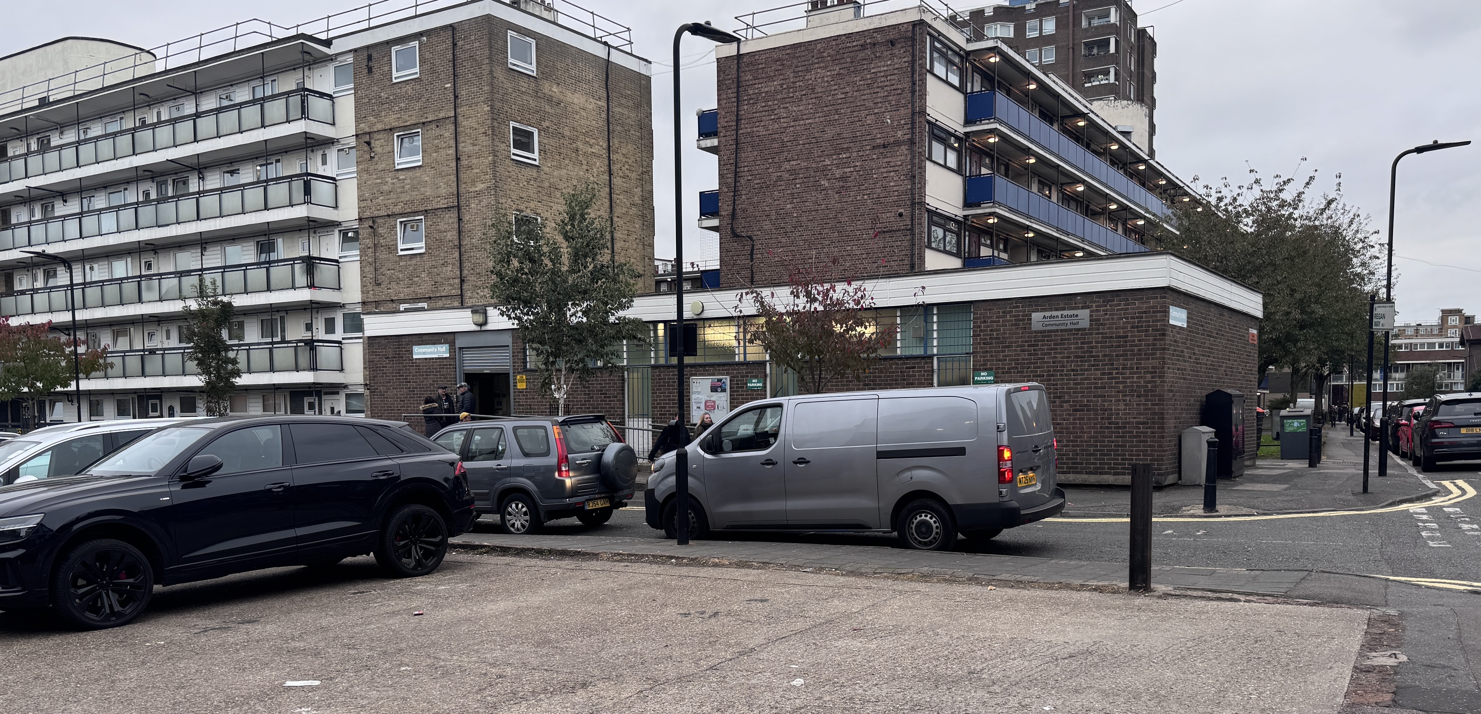Photograph of Arden Estate Community Hall viewed from the corner of Purcell Street and Regan Way. The single-storey brick community hall sits between residential blocks, with paved open space, parked vehicles and surrounding estate buildings visible.