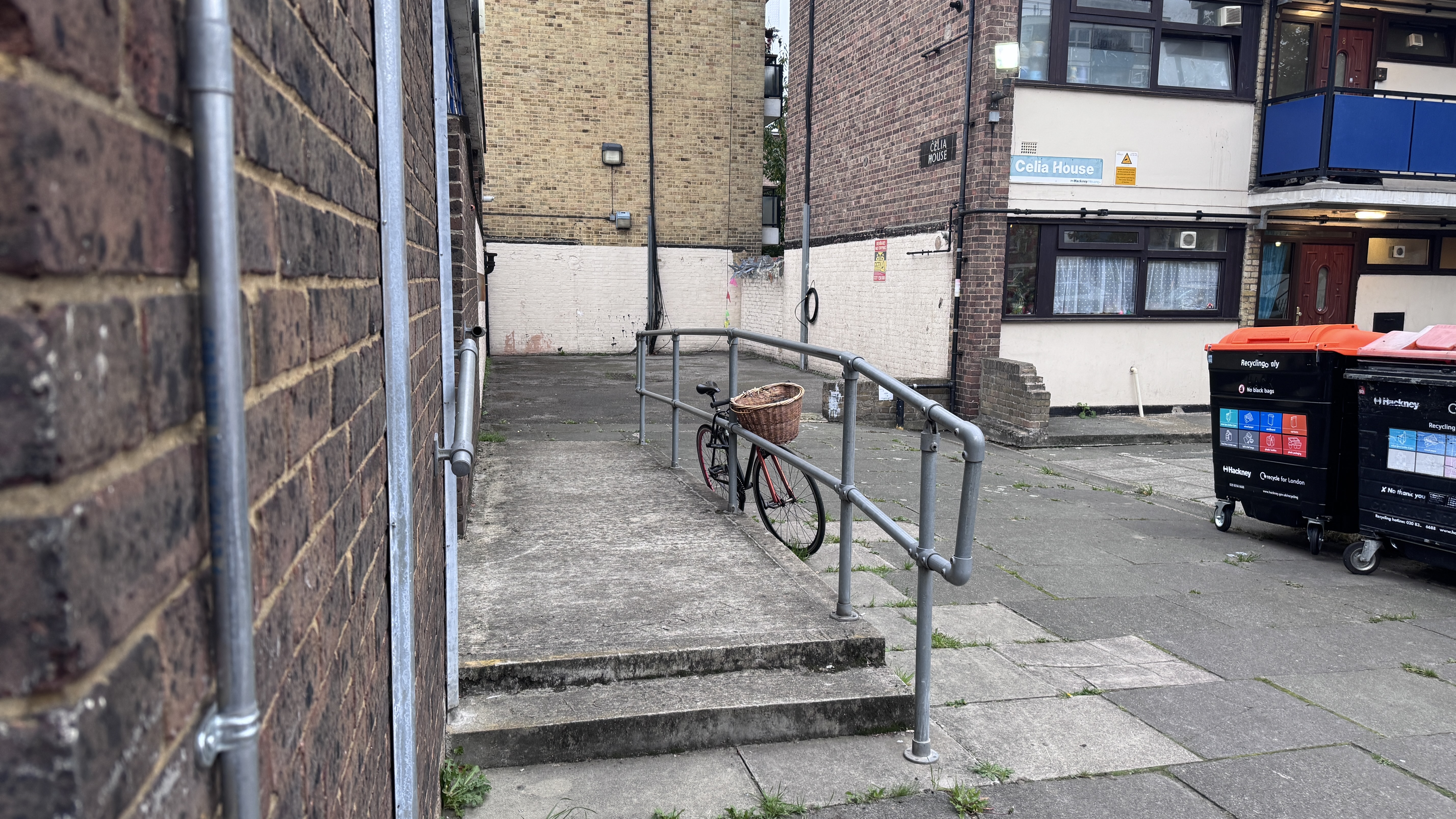 Photograph showing the space behind Arden Estate Community Hall between the hall and Celia House. The image shows a paved area with a wheelchair access ramp, handrails, a bicycle, refuse bins and surrounding residential buildings.