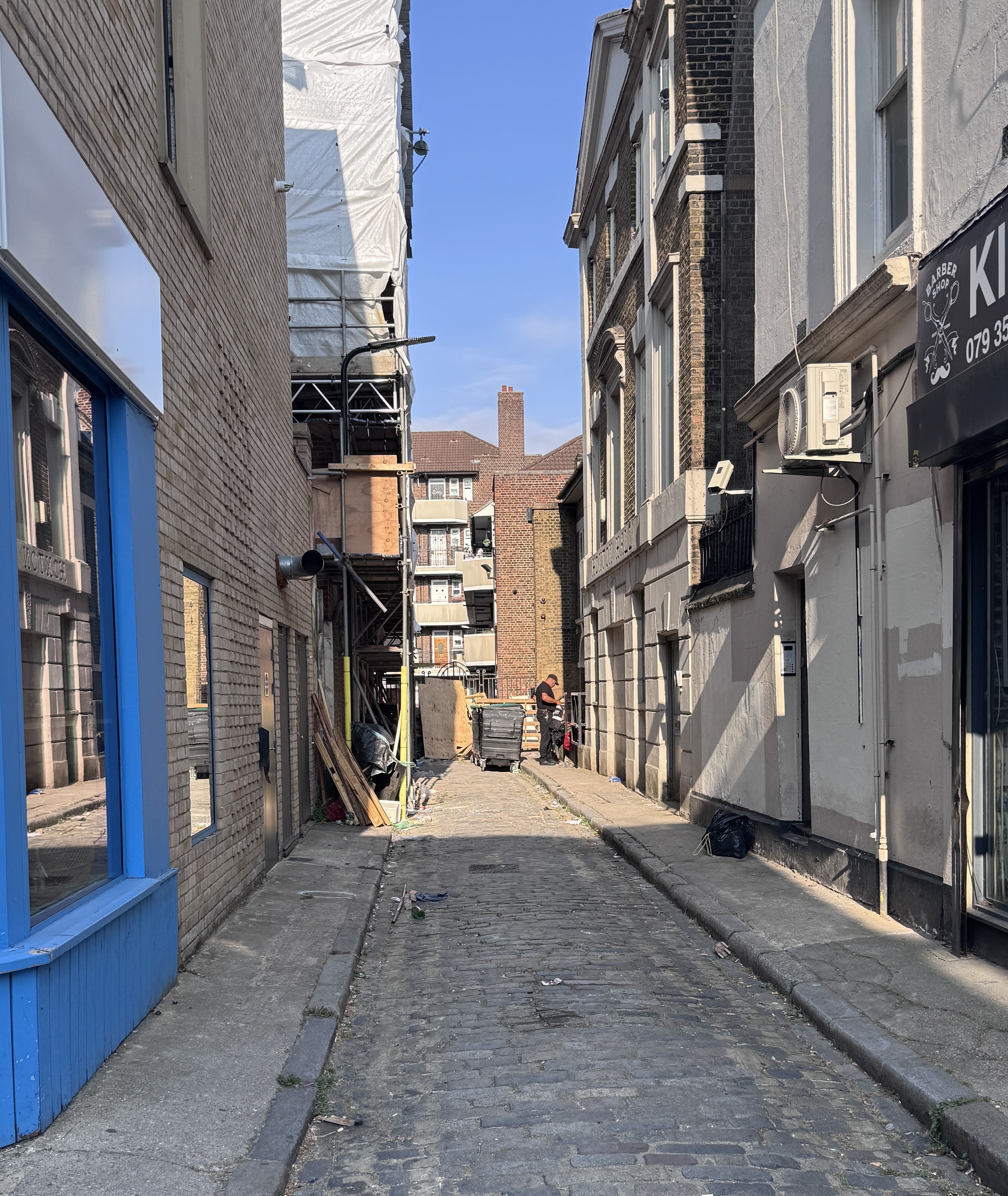 Photograph of Wilks Place, viewed from Hoxton Street. The cobbled lane runs between Hoxton Hall on the right and a building under construction on the left with scaffolding and protective sheeting. Construction materials are stored along the street, with residential buildings visible at the far end.